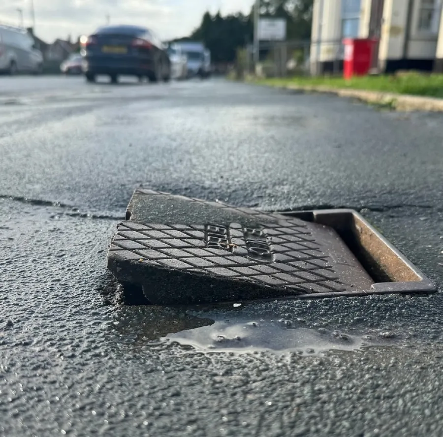 Close-up of a broken road vent cover partially lifted on an asphalt street with parked cars in background.