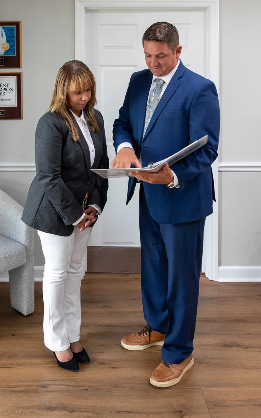 Man in blue suit explaining documents to woman in black blazer and white pants in office setting
