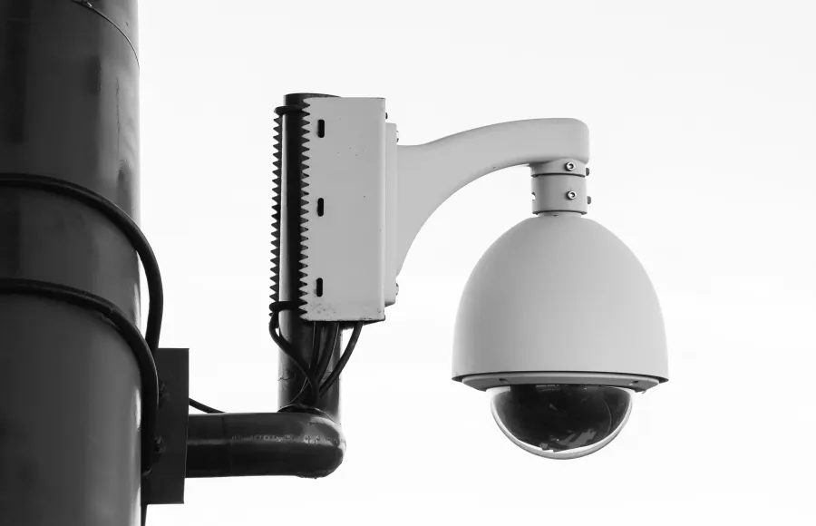 Black and white image of a dome security camera mounted on a pole against a bright sky.