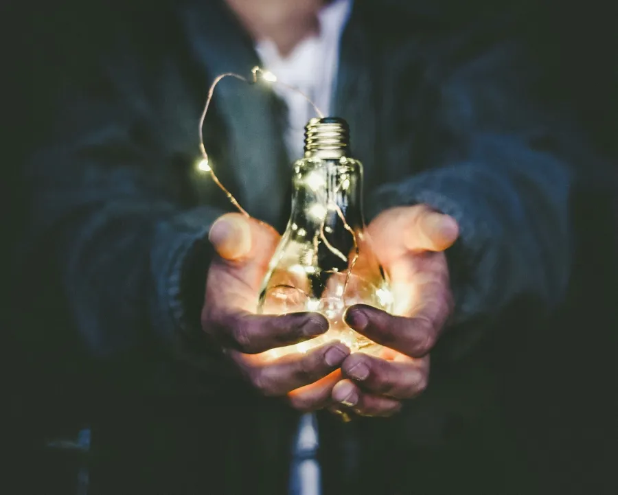 Person holding a glowing light bulb filled with string lights against a dark background.