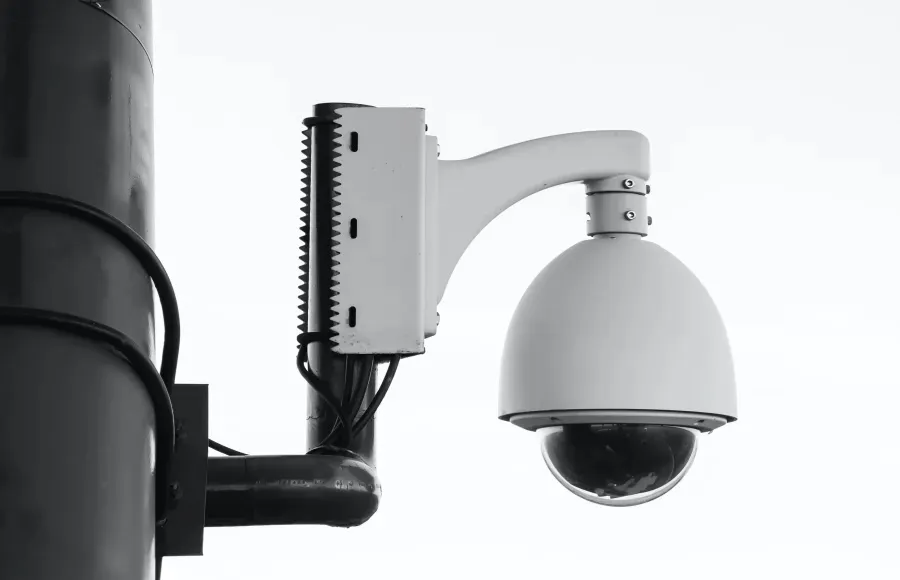 Black and white close-up of a dome security camera mounted on a pole against a bright sky.