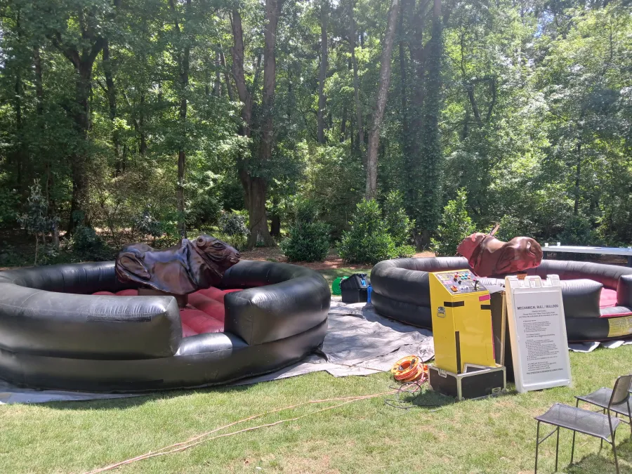 Outdoor mechanical bull ride setup with two inflatable arenas surrounded by trees in sunny weather