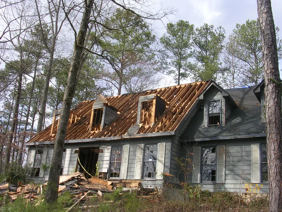 Old gray house with roof stripped down to wooden rafters surrounded by trees and debris on the ground