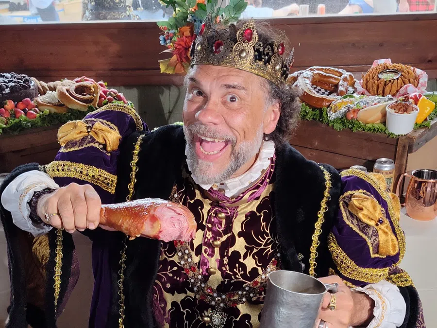 Smiling man in royal costume with jeweled crown holds turkey leg and metal mug at feast table.