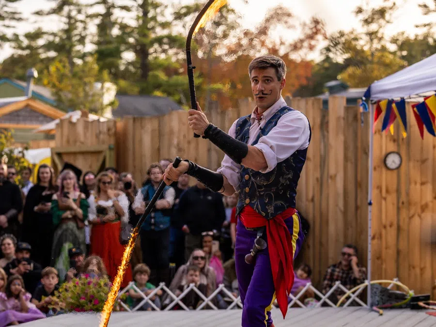 Fire performer in colorful medieval costume twirling a flaming whip on outdoor stage at festival with audience.