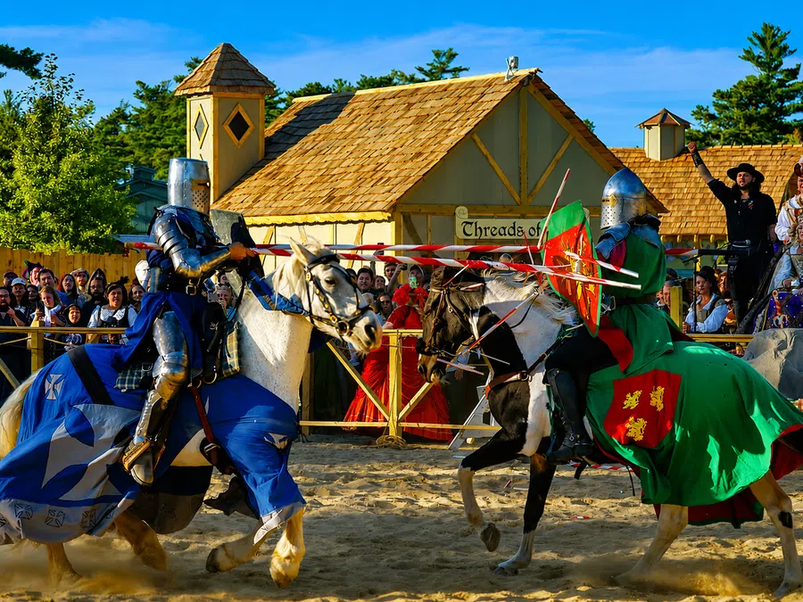 Two knights in armor jousting on horseback during a medieval festival with spectators and period buildings.