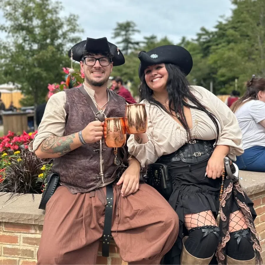 Smiling couple in pirate costumes holding copper mugs and sitting outdoors by flowers and brick wall.