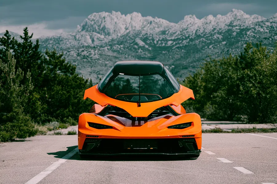Sleek orange sports car with butterfly doors open parked on mountain road under cloudy sky.