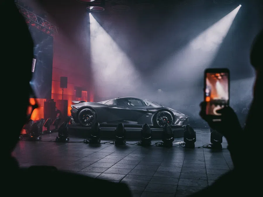 Sleek sports car displayed on stage with spotlights and audience capturing photo on smartphone.
