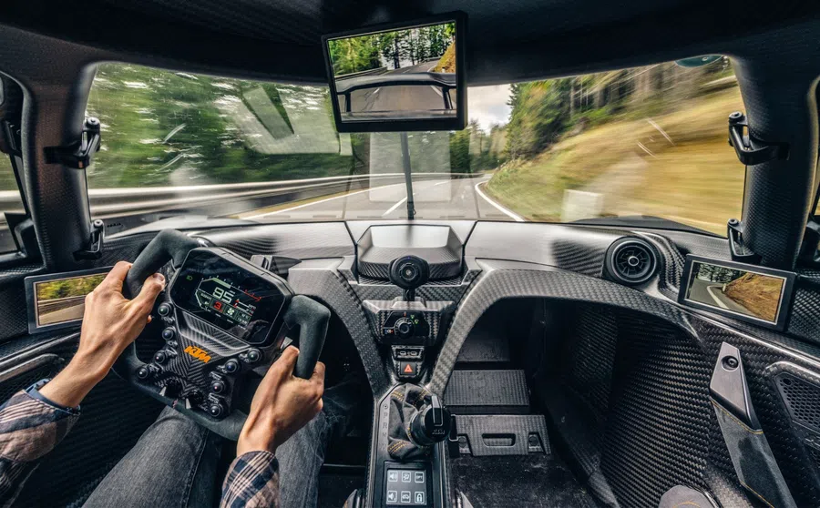 Driver view inside a high-tech race car cockpit with digital steering wheel and multiple cameras on a winding road.