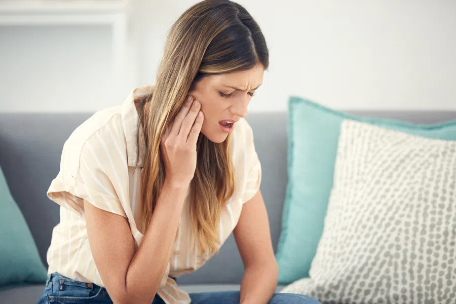 Woman sitting on couch holding her jaw in pain, showing signs of toothache or jaw discomfort indoors.