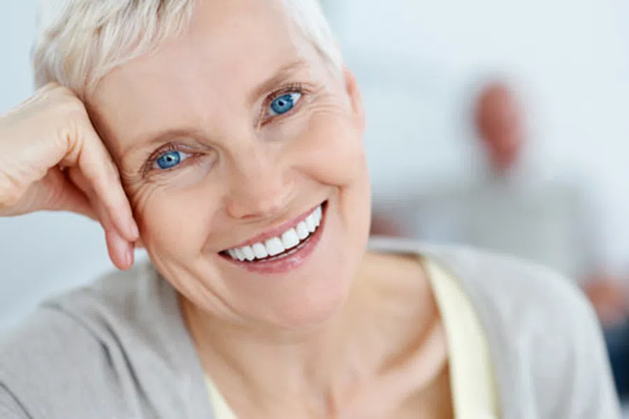 Close-up of a smiling elderly woman with short white hair and bright blue eyes in a soft-focus indoor setting.