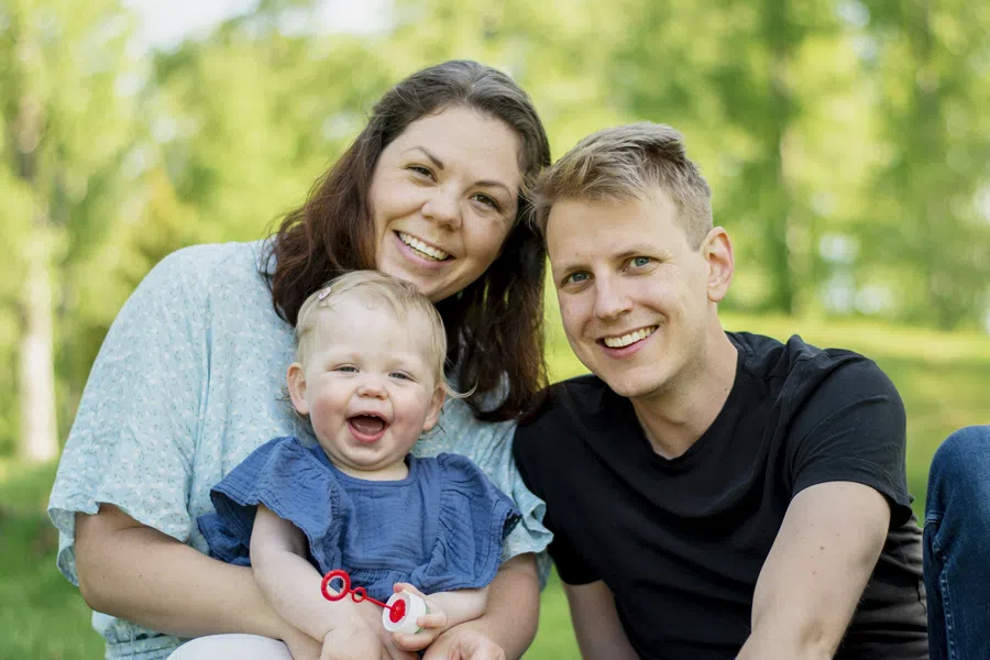 Smiling young family of mother, father, and baby sitting outdoors on grass with greenery background.