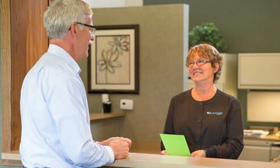 Smiling female receptionist handing a green card to an elderly male visitor at an office reception desk.