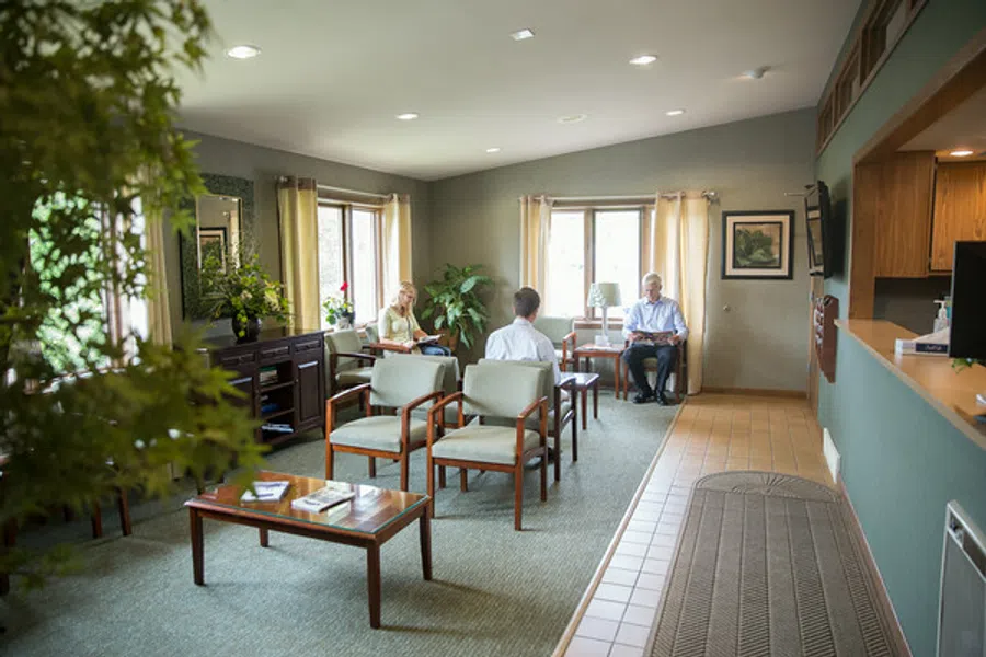 Bright waiting room with wooden chairs, tables, plants, and three people seated near windows.
