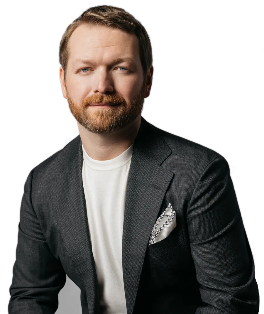 Man with red beard wearing a charcoal blazer and white shirt, posing with a neutral expression against transparent background.