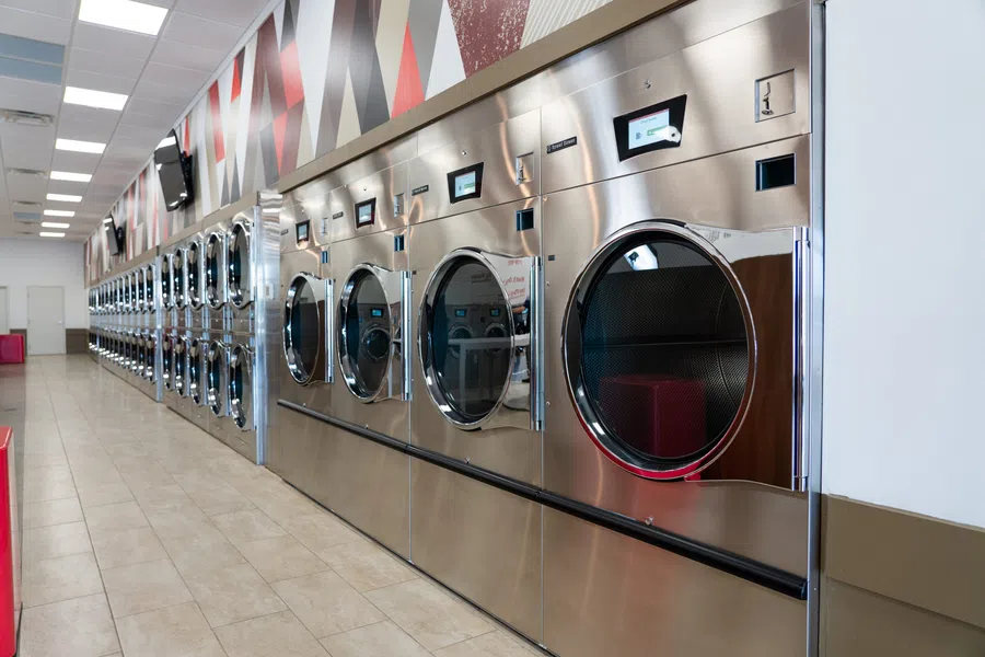 Row of stainless steel industrial washers with digital displays in a brightly lit laundry facility.