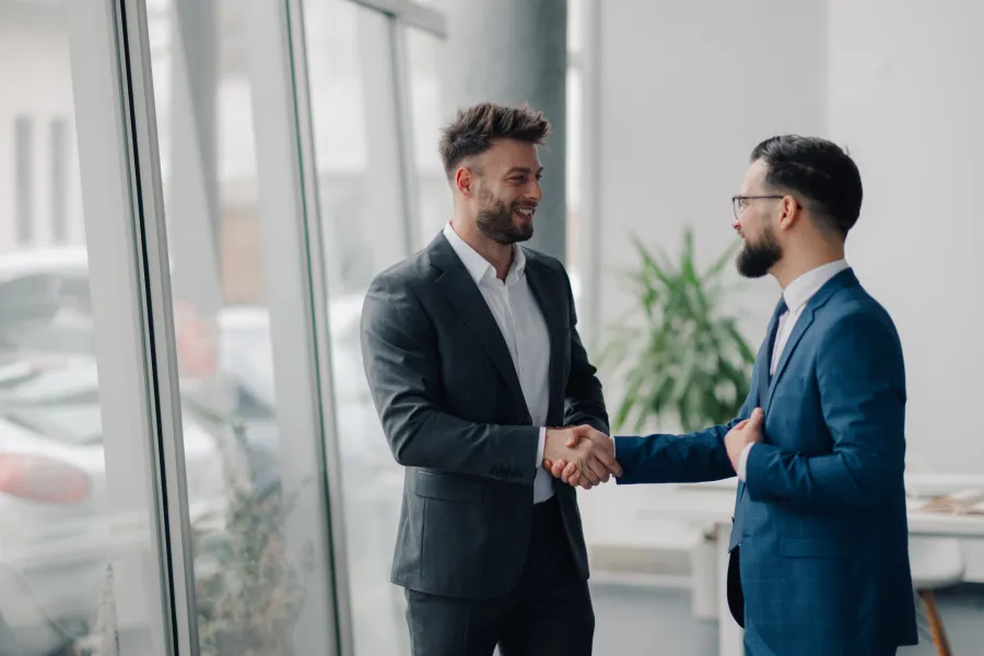 Two businessmen in suits shaking hands in a modern office with large windows and plants