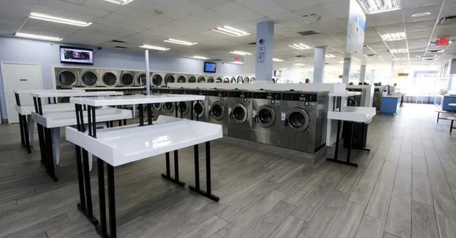 Bright, clean laundromat with rows of washers and dryers and folding tables on wood-style tile flooring