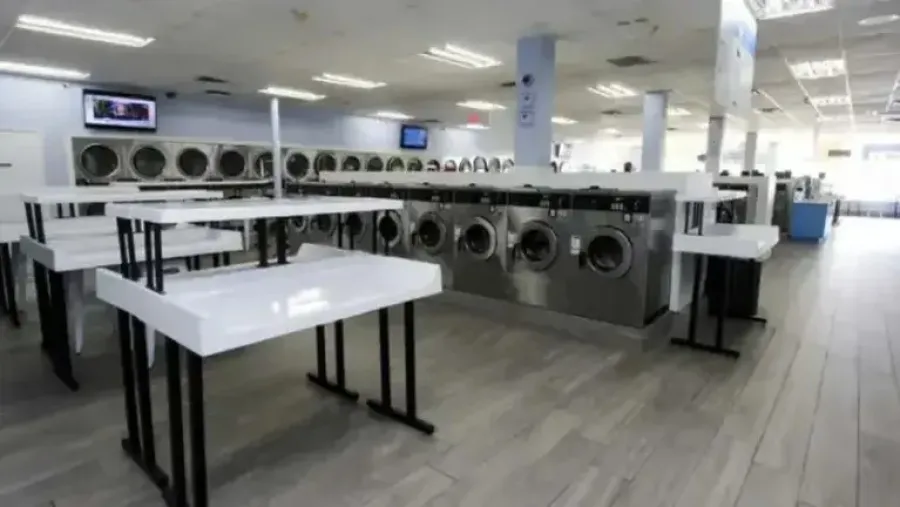 Bright, spacious laundromat with rows of stainless steel washing machines and white folding tables on light wood flooring.