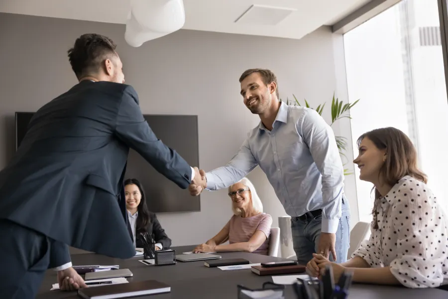Two men shaking hands in a modern office meeting with three colleagues observing and smiling.