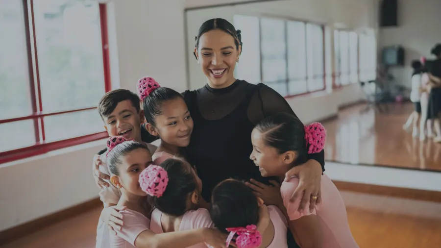 Ballet teacher embraces smiling young dancers in a studio with wooden floors and large windows.