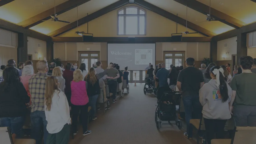 People standing inside a church auditorium facing a welcome screen during a service or event.