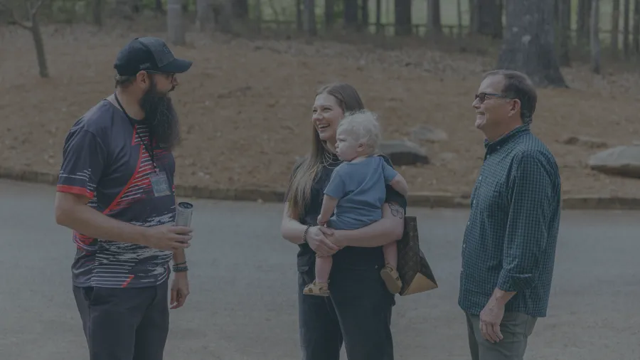 Group of three adults and one child outdoors chatting on a paved area near a wooded park.