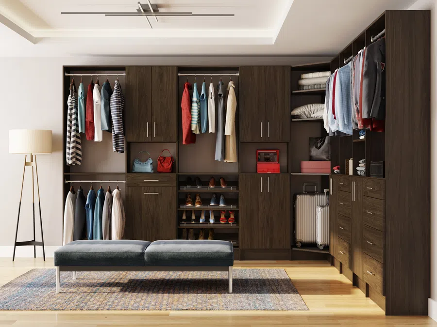 Modern walk-in closet with dark wood cabinets, hanging clothes, shoe racks, and a padded bench on rug.