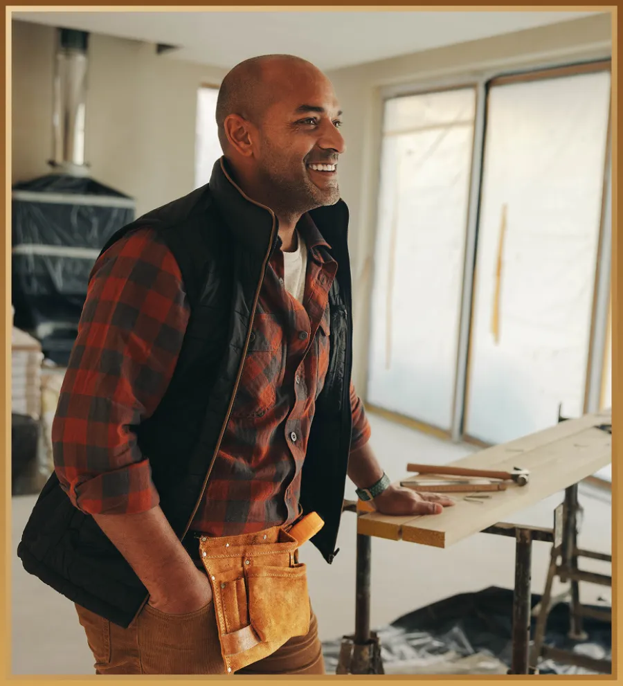Smiling male carpenter in plaid shirt and tool belt leaning on workbench with tools in a workshop under renovation