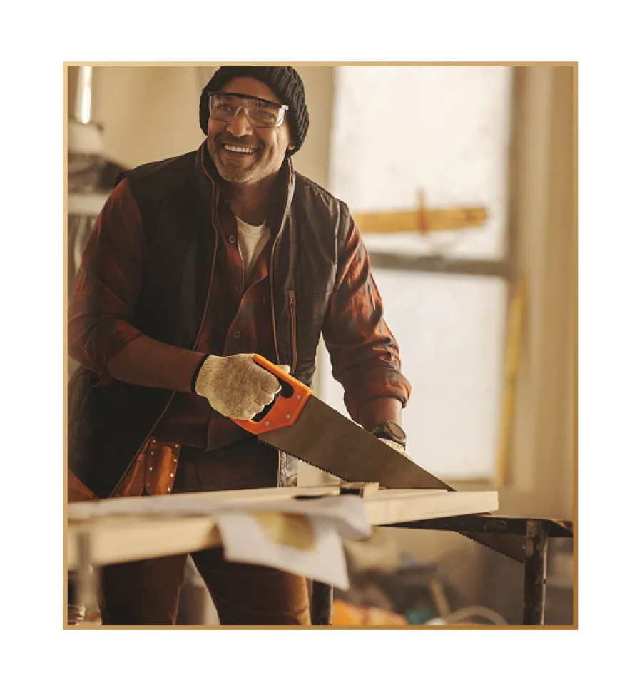 Smiling man wearing protective glasses and gloves sawing wood indoors in a bright woodworking studio.