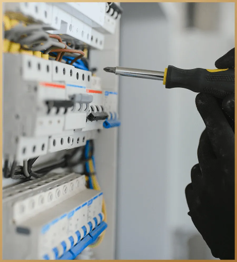 Hand in black glove using screwdriver to adjust circuit breaker panel with colored wires and switches.