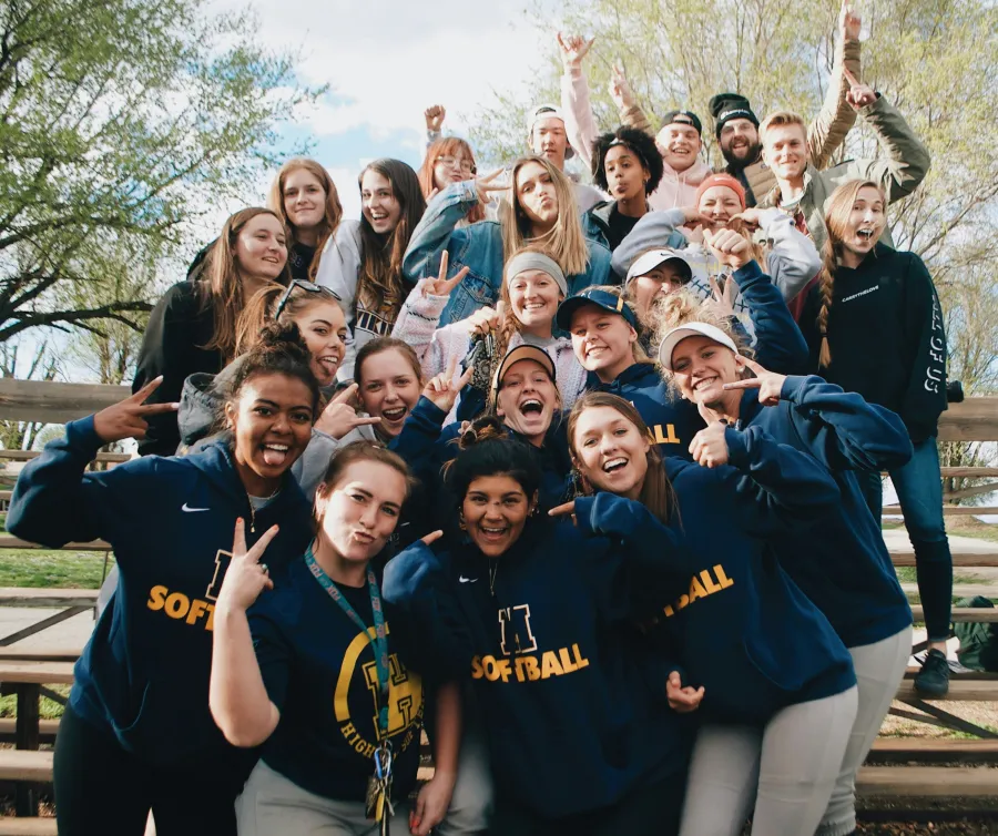 Happy group of young softball players and friends posing energetically outdoors on wooden bleachers.