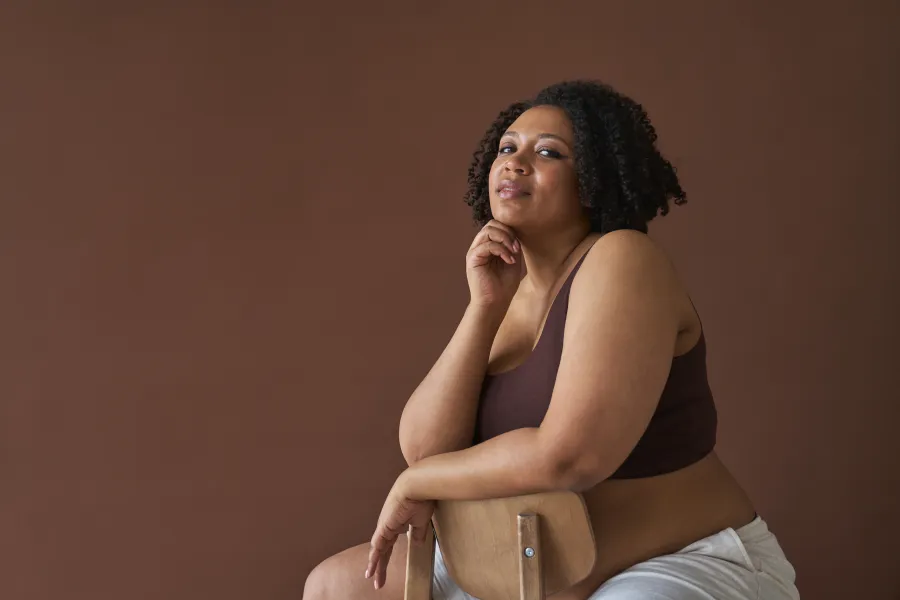 Confident woman with natural curly hair wearing a brown tank top sitting on a wooden chair against a brown background.