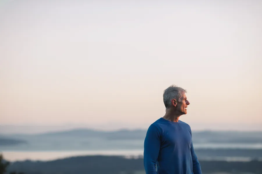 Side view of a mature man in a blue sweater looking at a serene, foggy lake landscape during sunrise or sunset.