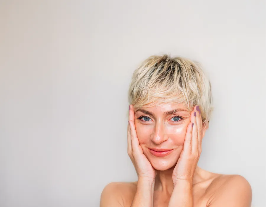 Smiling woman with short blonde hair gently touching her face against a neutral background.