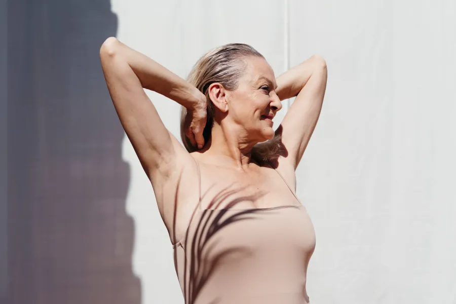 Smiling older woman stretching arms behind head wearing a sleeveless beige top with shadow on skin.