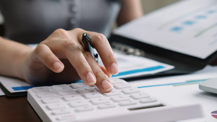 Person using a white calculator with financial documents and charts on a desk during work.
