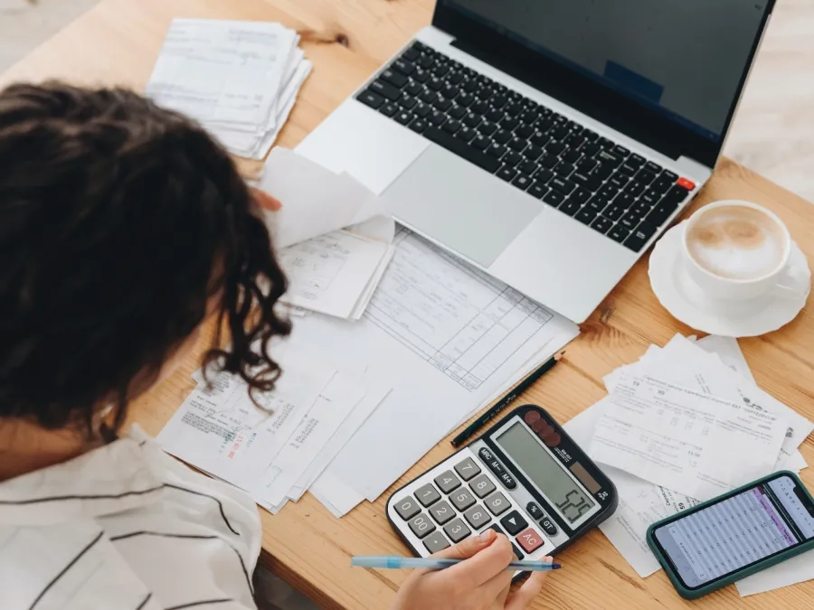 Person using calculator and laptop to review bills and expenses at a wooden desk with coffee and phone nearby