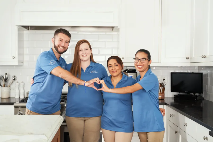 Four smiling people in blue shirts form a heart shape with their hands in a bright white kitchen.