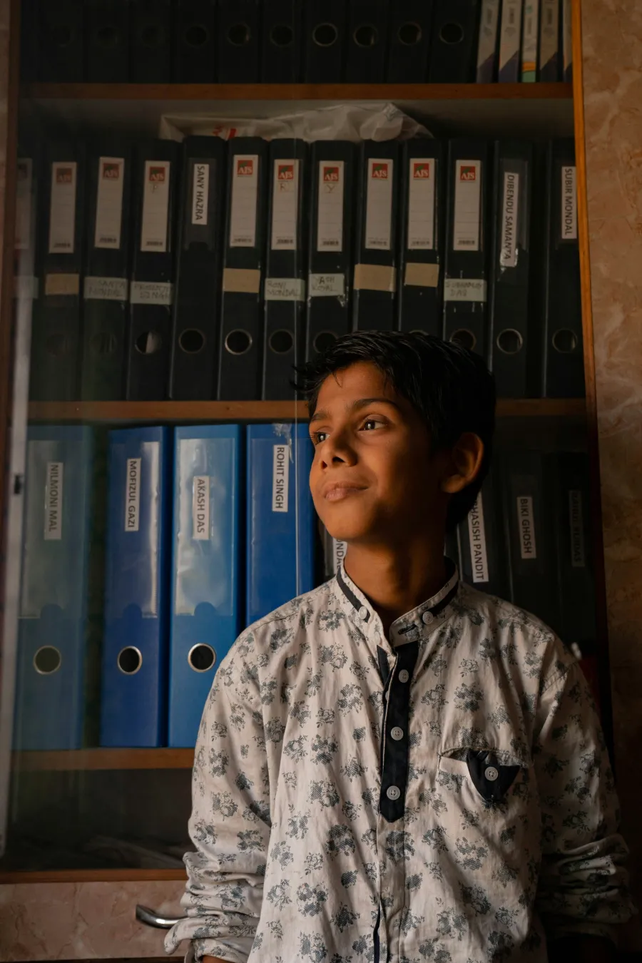 Young boy in a patterned shirt stands thoughtfully in front of a shelf filled with labeled black and blue binders.