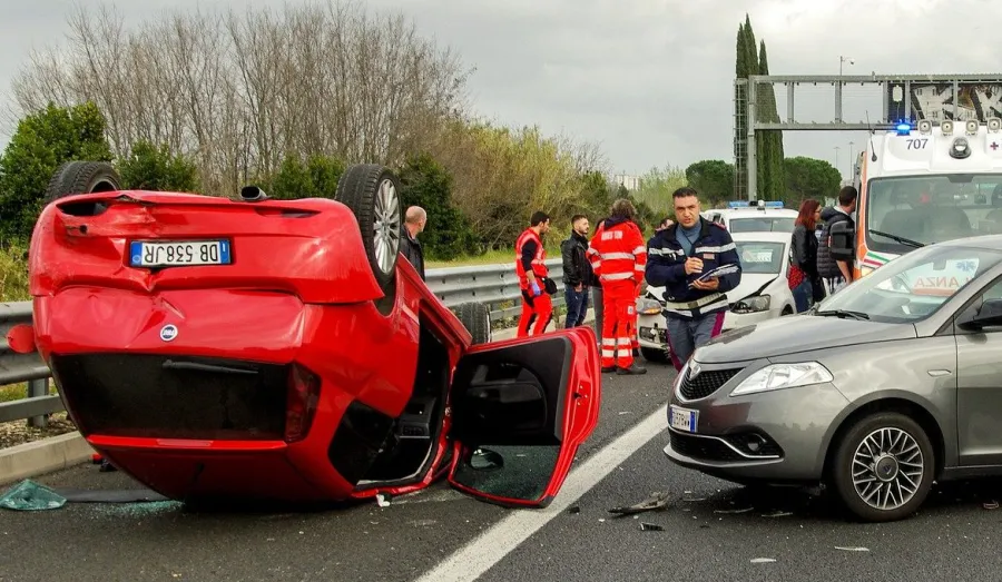 Overturned red car in a highway accident with emergency responders and a gray car nearby on cloudy day