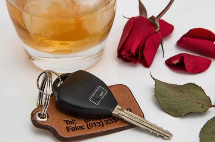 Car key with leather keychain beside a glass of whiskey and red rose on a white surface