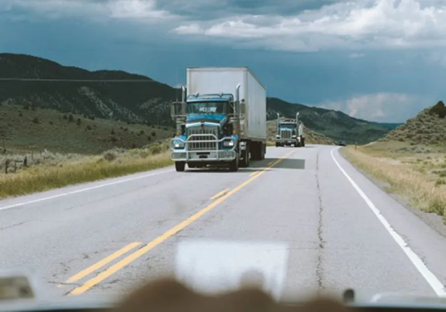 Two blue semi trucks driving on a rural highway surrounded by green hills under a cloudy sky.