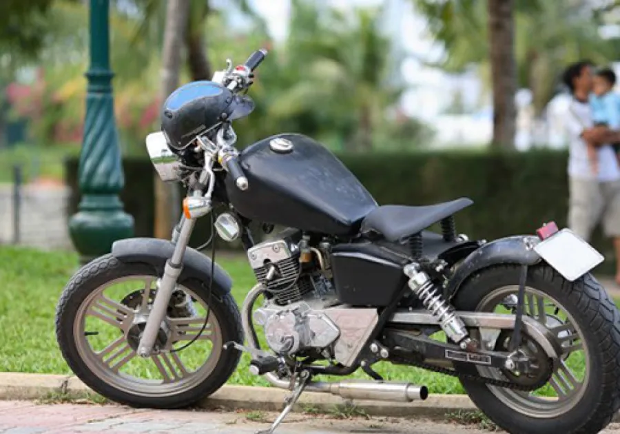 Black classic motorcycle parked on pavement with green park and blurred people in background