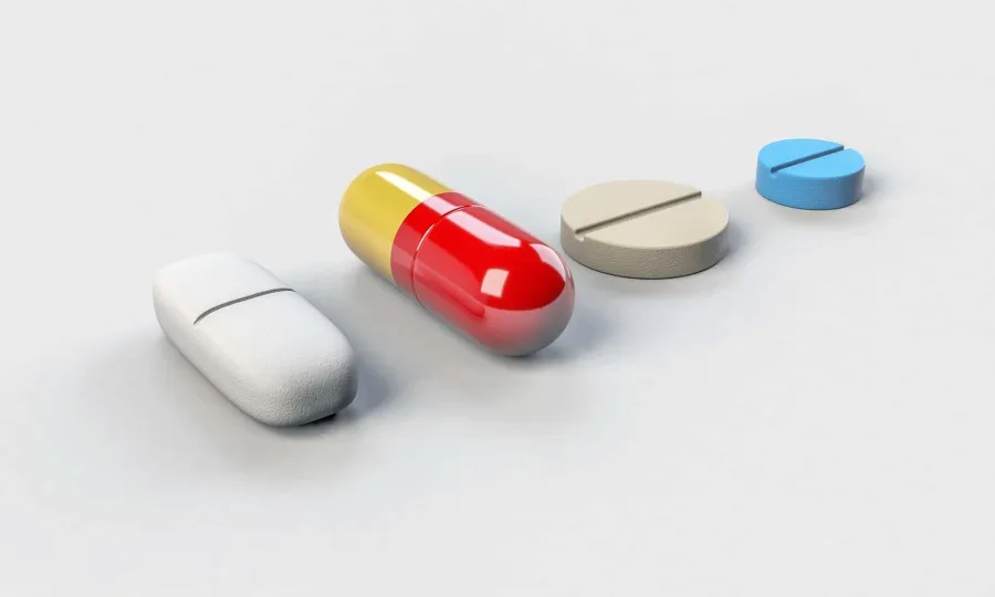 Various medications including a white capsule, red and yellow pill, beige and blue tablets on a white surface.