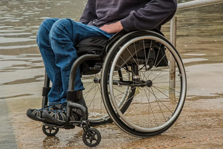 Close-up of a person in a wheelchair near a calm water body, wearing blue jeans and a dark sweatshirt.