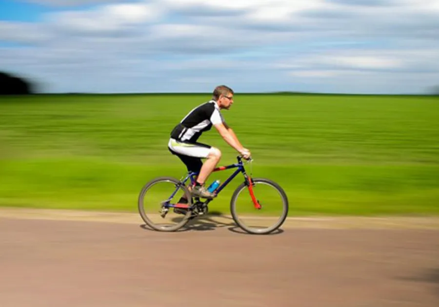 Man wearing black and white cycling gear riding a mountain bike on a road with green fields and a blue sky background