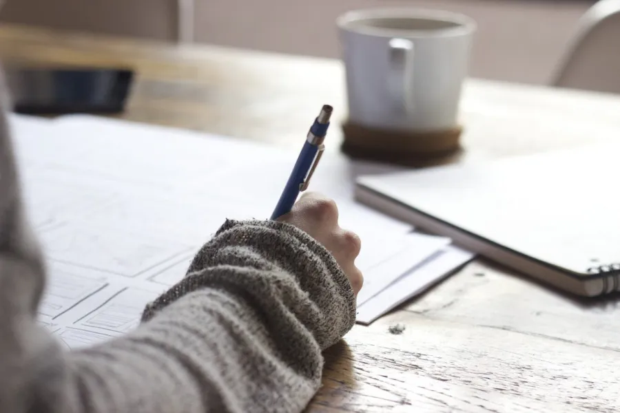 Person writing on paper with blue pen at wooden table, coffee cup and notebook nearby in cozy setting