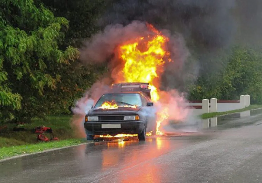 Car engulfed in flames on a rainy road near green trees and a white guardrail.